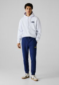 Weißer Hoodie mit einer Fronttasche und einem Logo, kombiniert mit marineblauen Jogginghosen mit gelbem Text, abgerundet mit lässigen Turnschuhen.