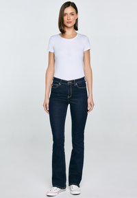 Egyenes, sötét hajú nő, fehér rövid ujjú pólót, sötétkék, magasderekú farmert és fehér sportcipőt visel, szemből áll.