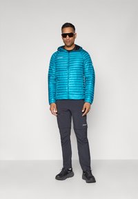 Blaue gesteppte Daunenjacke mit Reißverschluss und Kapuze, kombiniert mit schwarzen Reißverschluss-Cargo-Hosen und dunklen Sneakers. Das Modell trägt dunkle Sonnenbrillen.