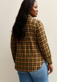 Camisa a cuadros marrón hecha de tela ligera, con patrones de cuadros en negro y amarillo, mangas largas y dobladillo curvo.