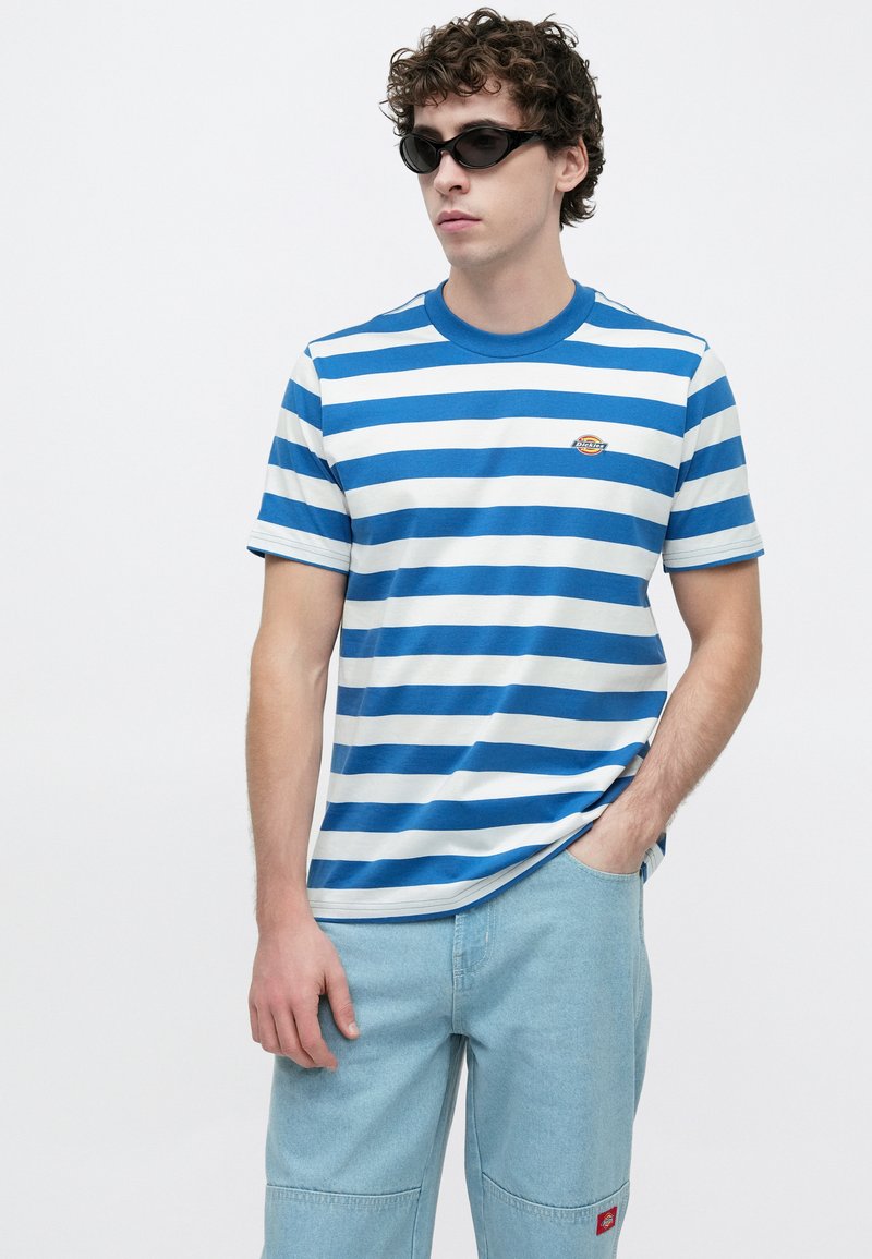 Maglietta a righe blu e bianche in cotone con scollo rotondo. Presenta maniche corte e un piccolo logo sul petto. Abbinata a pantaloni azzurri.