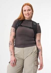 Graues, eng anliegendes Hemd, Cargo-Hosen in hellbeige, schwarzer Rucksack mit Riemen. Tattoos an den Armen sichtbar, Zubehör umfasst Ringe. Heller Hintergrund.