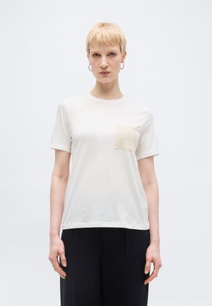 Osoba o krótkich blond włosach, ubrana w białą koszulkę z teksturowaną kieszenią na lewej piersi i czarne spodnie, stojąca na jednolitym tle.