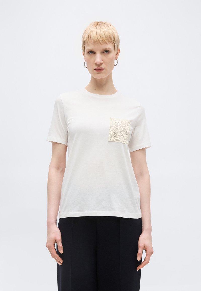 Άτομο με κοντάξανθά μαλλιά που φοράει λευκό T-shirt με υφασμένη τσέπη στο αριστερό στήθος και μαύρο παντελόνι, στέκεται μπροστά σε απλό φόντο.