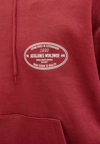 Haut à capuche bordeaux avec un logo en texte blanc sur la poitrine indiquant "Établi à Copenhague en 1990 Jack&Jones Mondial De la Vision à la Réalité."
