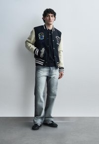 Νέος άνδρας που φοράει μαύρο και λευκό varsity μπουφάν, μαύρο πουκάμισο, ξεθωριασμένο γκρι τζιν και μαύρα αθλητικά παπούτσια, στέκεται μπροστά σε έναν απλό τοίχο.