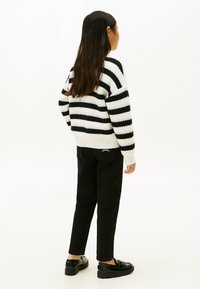 Jongere met lang zwart haar, gekleed in een zwart-wit gestreepte trui, zwarte broek en zwarte instappers, staand met de rug naar ons gekeerd.