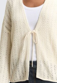 Gebreid crèmekleurig vest met ajourpatroon, voorgebonden, gedragen over een witte geribbelde tanktop en donkere spijkerbroek.