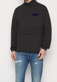 Černý pulovr na zip s vysokým límcem, má přední kapsu, elastické manžety a texturovanou výšivku na hrudníku. Nosený s popranými džíny.
