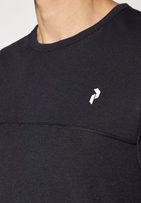 Nahaufnahme einer Person, die ein schwarzes T-Shirt mit einer horizontalen Naht und einem kleinen weißen geometrischen Logo auf der Brust trägt.