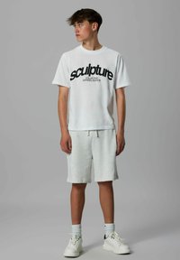 T-shirt blanche avec le texte noir "sculpture COLLECTIVE APPAREL EDITION," associée à un short gris clair et des baskets blanches avec des accents noirs.