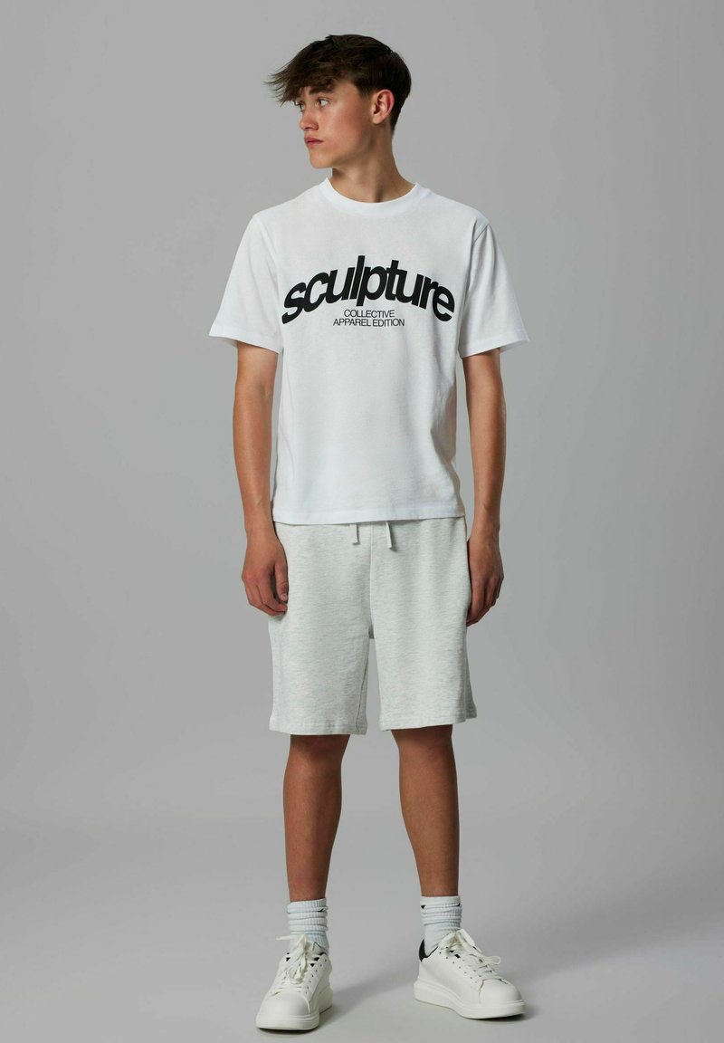 T-shirt blanche avec le texte noir "sculpture COLLECTIVE APPAREL EDITION," associée à un short gris clair et des baskets blanches avec des accents noirs.