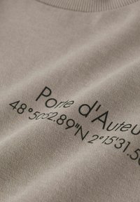 Lyse beige sweatshirt med sort tekst, der lyder "Porte d'Auteur 48°50'28.89"N 2°15'31.5"E." Glat tekstur, ribbet halsudskæring.