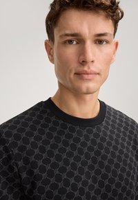 Czarna bluza z geometrycznym wzorem w odcieniach szarości. Posiada okrągły dekolt i prążkowane wykończenia. Materiał wydaje się miękki i strukturalny.