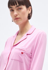 Set de pijamale roz, cu un buzunar la piept decorat cu o inimă roșie și cu borduri roșii contrastante pe guler și pe fanta nasturilor. Material moale și fin.