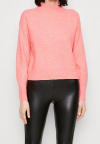 Růžový pletený svetr s vysokým límcem a žebrovaným lemem, s dlouhými, volnými rukávy. Kombinován s černými kalhotami vzhledem k pokožce.