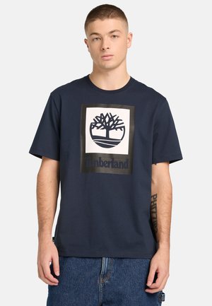 Junger Mann mit kurzen Haaren, der ein marineblaues Timberland-T-Shirt und blaue Jeans trägt und vor einem schlichten hellen Hintergrund steht.