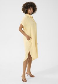 Abito giallo con bottoni, maniche corte, tasche laterali e uno spacco alto. Realizzato in tessuto testurizzato, presenta una vestibilità comoda e un colletto.