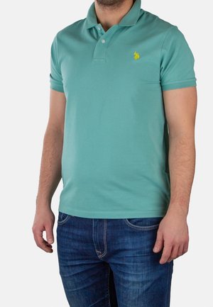 Mężczyzna w turkusowej koszulce polo z krótkim rękawem z żółtym logo na piersi oraz niebieskich dżinsach, stojący na jednolitym tle.