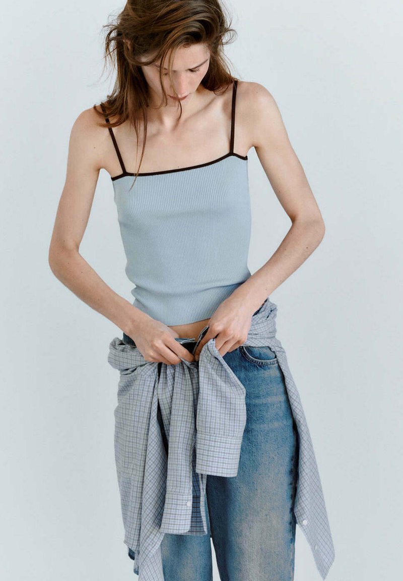 Femme aux longs cheveux portant un débardeur bleu clair et un jean, attachant une chemise à carreaux bleue nouée autour de sa taille.