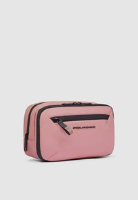 Organizador de viaje rosa con textura suave, detalles en cremallera negra y un bolsillo frontal. Incluye un logo en el lateral en color negro. Forma rectangular.