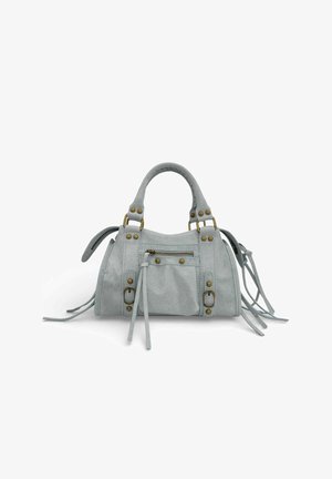 Sac à main en daim gris clair avec deux poignées, poche zippée à l'avant, boucles décoratives, clous en laiton et longs accents de pompons sur fond blanc.