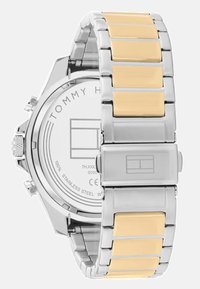 Silberne Edelstahl-Uhr mit goldenen Akzenten am Armband. Verfügt über einen strukturierten Rücken und einen rechteckigen Verschluss. Logo auf der Rückseite sichtbar.
