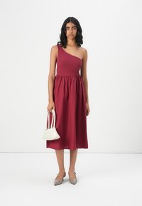 Robe bordeaux à une épaule, avec un corsage ajusté et une taille froncée. Le mannequin tient un petit sac à main blanc, associé à des chaussures beiges à bouts pointus.