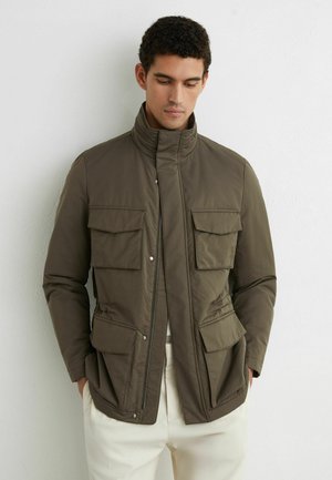 Mladý muž oblečený v olivovo zelenej bunde so štyrmi prednými klopnými vreckami, stojaci opretý o bielu stenu, ruky vreckách, pozerajúci sa dole.