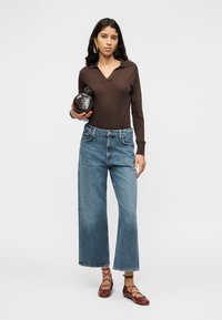 Rudas ilgomis rankovėmis megztinis su apykakle, mėlyni plačios kelnės ir tamsiai raudoni lygūs batai. Modelis laiko juodą apvalią rankinę.