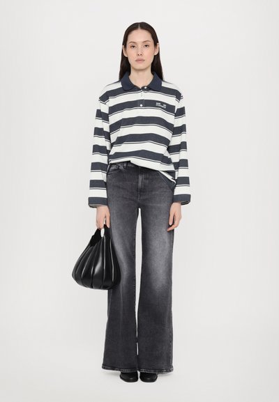Langärmliges Polohemd mit navyblauen und weißen Streifen, kombiniert mit weit geschnittenen grauen Jeans. Hält eine schwarze Faltenhandtasche. Vor einem weißen Hintergrund.