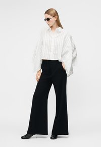 Žena oblečená v čiernych širokých nohaviciach, bielej voľnej košeli na gombíky, čiernych slnečných okuliaroch a čiernych lodičkách, stojaca s jednou rukou v kapse.