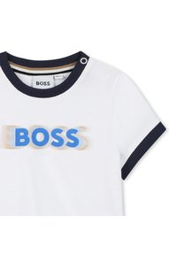 Bílé tričko s námořnicky modrým žebrovaným límcem a manžetami na rukávech. Obsahuje velké modré logo "BOSS" s rozostřeným efektem. Dva patenty na ramenou.