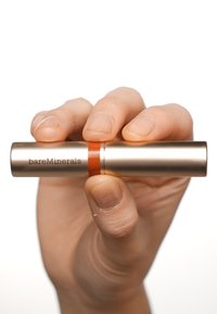 Prodotto per labbra BareMinerals in un tubo cilindrico dorato, con una banda di accento arancione. Tenuto in mano, che mette in mostra una finitura liscia e metallica.
