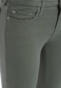 Jeans elásticos de color verde oliva con una textura suave, que cuentan con un bolsillo frontal y un pequeño detalle de logo metálico en el lateral.