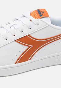 Sneakers bianchi con un accento arancione, caratterizzati da una tomaia sintetica liscia, una scatola per le dita perforata e lacci bianchi piatti.