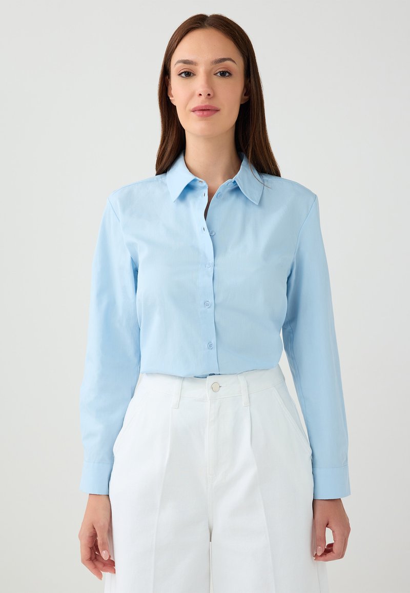 Light blue button-up shirt with long sleeves, a collared neckline, and smooth texture, paired with high-waisted white trousers.