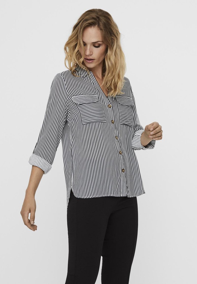 Blouse rayée noir et blanc avec un bouton sur le devant, deux poches poitrine et des manches retroussées, fabriquée en tissu léger. Coupe ajustée.