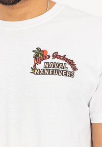 Hvid bomulds t-shirt med et farverigt trykt design, hvor "Alpha Industries" står over "NAVAL MANEUVERS" med palme-aksenter.