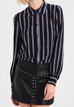 Donna che indossa una camicia a righe verticali nere, blu e bianche con bottoni e una gonna in pelle nera con dettagli in borchie argento.
