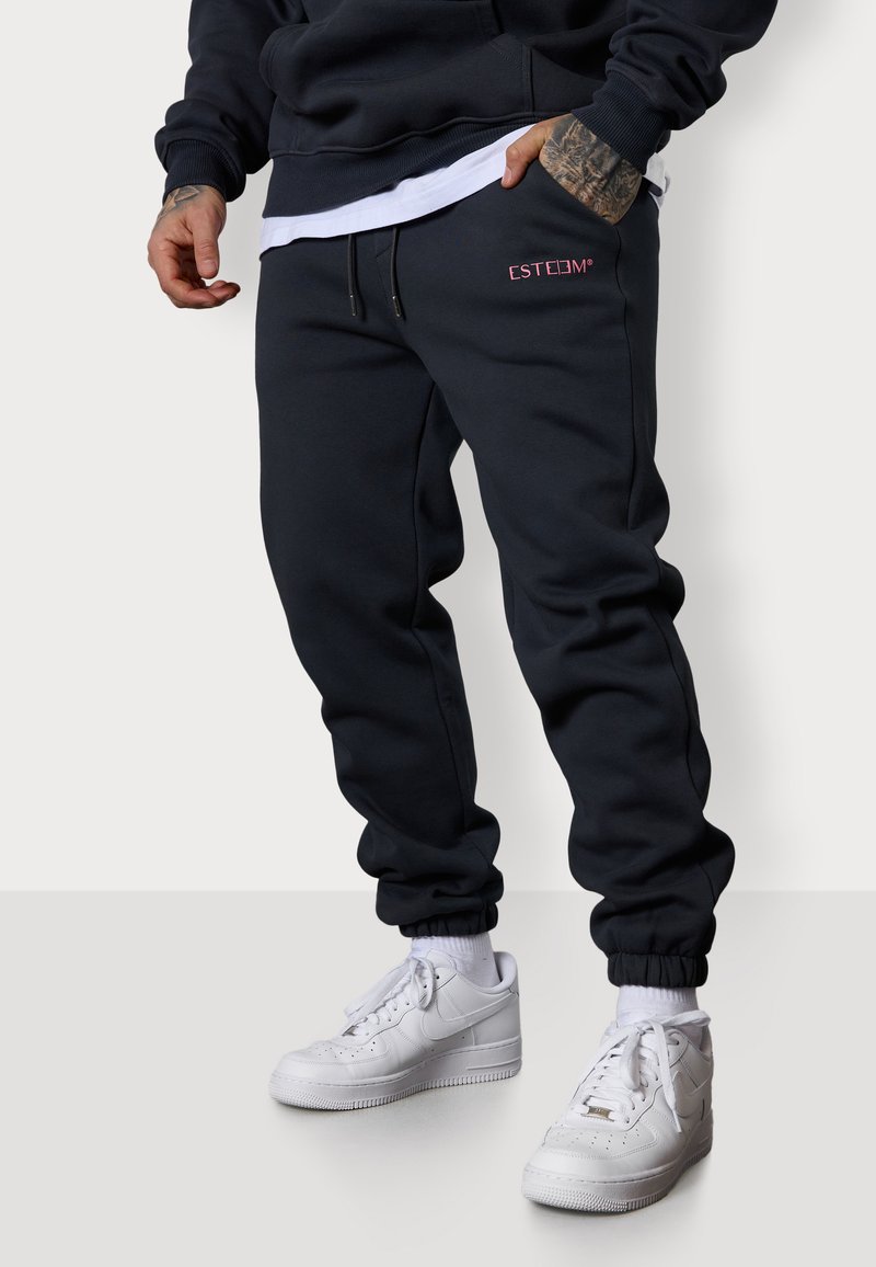 Dunkelblaue Sweatpants mit elastischen Bündchen und Seitentaschen; mit pinkem gesticktem Logo. Kombiniert mit weißen Sneakers. Glatte Stofftextur.