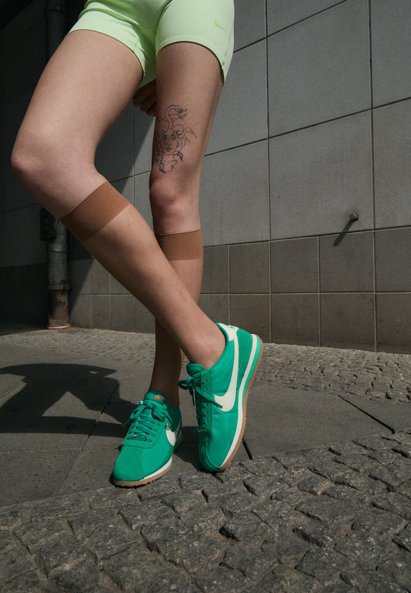 Zapatillas Nike turquesas con swoosh blanco, combinadas con pantalones cortos verde lima. Fajas marrones en las rodillas y un tatuaje detallado en el muslo. Entorno urbano.