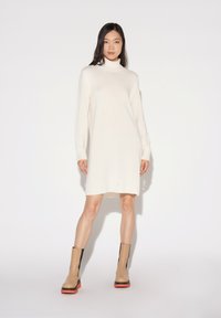 Abito lavorato a maglia color crema con colletto alto e maniche lunghe, con dettagli a coste sull'orlo. Indossato con stivali con tacco beige e neri.