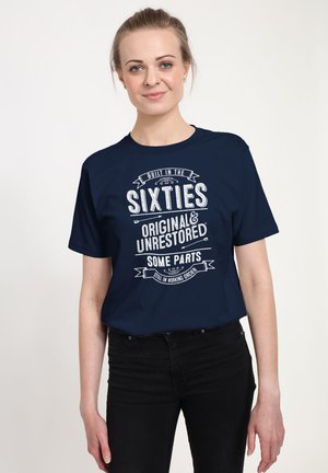 Femme aux cheveux blonds attachés, portant un t-shirt bleu marine avec l'inscription « Construit dans les années soixante Original et non restauré Certaines pièces encore en état de marche ».