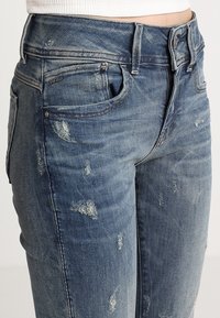 Jeans de mezclilla desgastados de color azul claro con un diseño ajustado, que presentan desgastes y roturas, cinco bolsillos y un cierre de botón.
