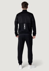 Completo sportivo nero con parte superiore posteriore testurizzata, pantaloni abbinati e sneakers bianche. Presenta loghi e grafiche sulla schiena.