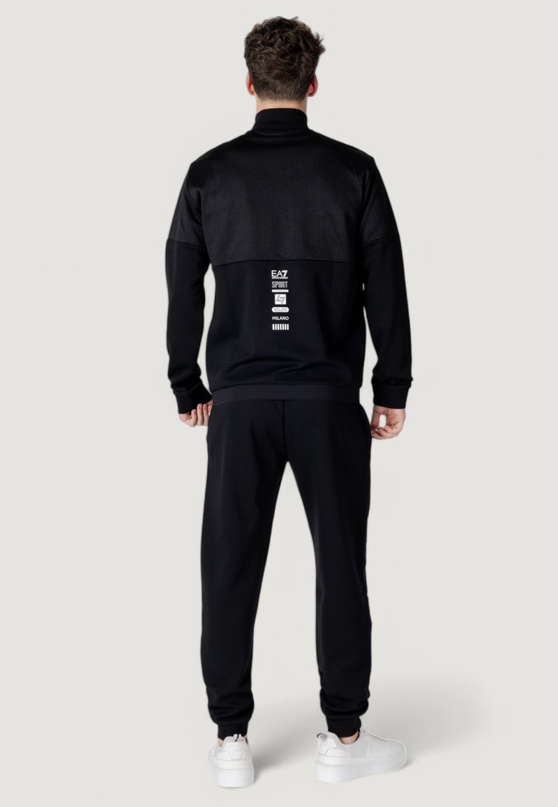 Completo sportivo nero con parte superiore posteriore testurizzata, pantaloni abbinati e sneakers bianche. Presenta loghi e grafiche sulla schiena.