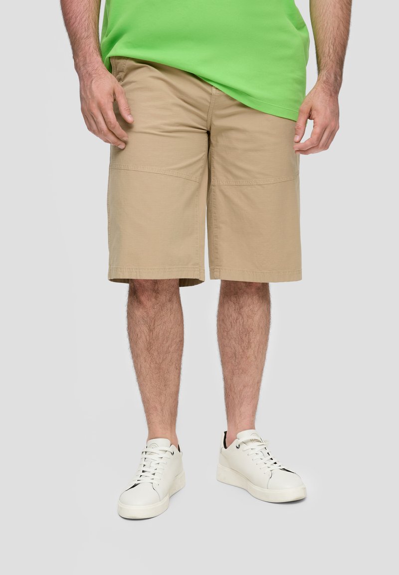 Mann in beigen knielangen Shorts, weißen Sneakern und einem grünen Hemd vor einem schlichten hellen Hintergrund.