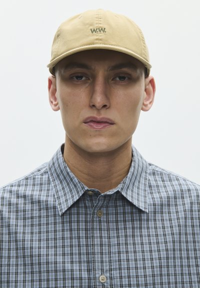 Nuori henkilö, jolla on beige baseball-lippis, jossa lukee "W.W.", ja sininen ruudullinen nappipaita, katsoo eteenpäin neutraalilla ilmeellä vaaleaa taustaa vasten.