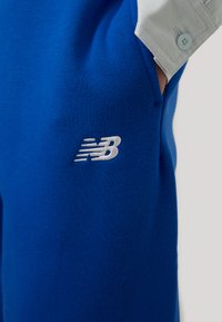 Zili sporta šorti, izgatavoti no mīksta auduma, ar izšūtu baltu New Balance logotipu sānos un gaiši pelēku kabatas detaļu.